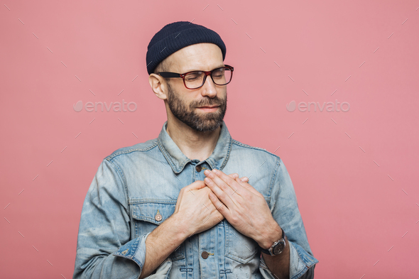 Friendly looking bearded male with closed eyes, keeps hands on chest, wears denim jacket