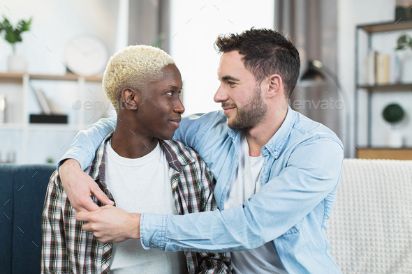 Multiracial gay couple sitting in embrace on comfy couch