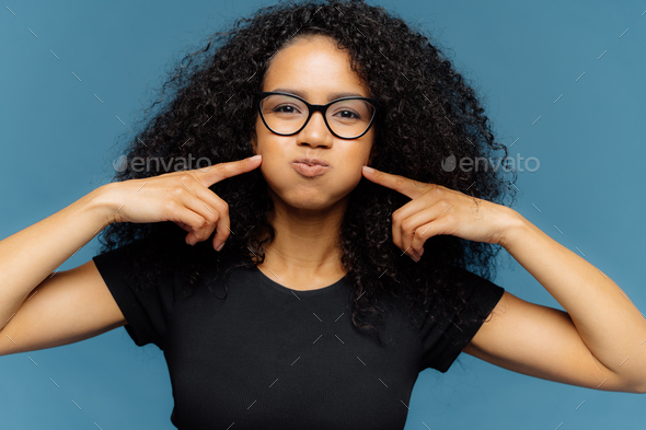 Funny dark skinned woman with curly hair, presses cheeks, holds breath, blows cheeks