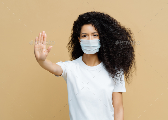 Woman makes stop gesture, pulls palm towards camera, wears medical flu mask