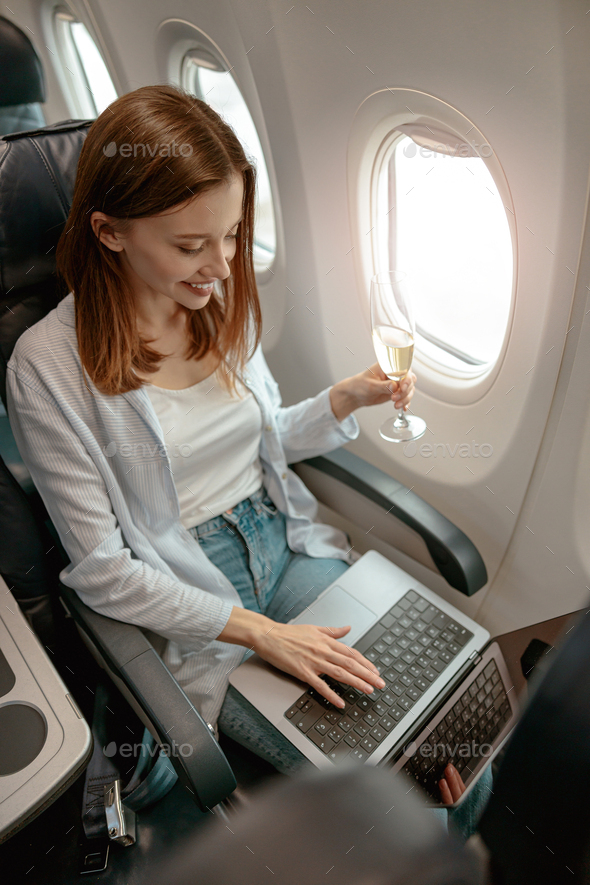 Joyful woman using laptop and drinking champagne in plane