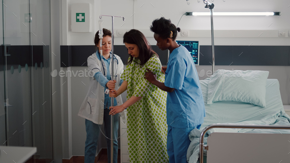Medical team helping patient to stand up from bed holding intravenous IV fluid drip bag