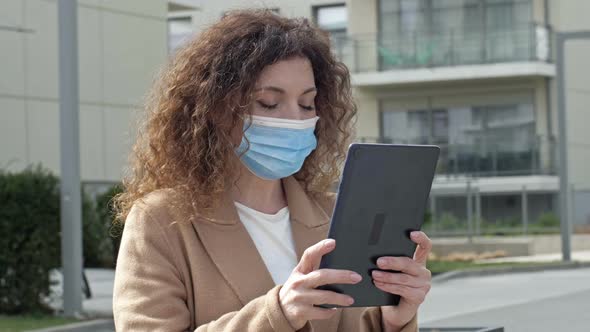 Woman in a Protective Mask is Standing in a Deserted Street and Chatting with Someone Using a Tablet