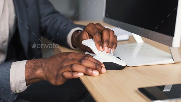 Unrecognizable black office worker wiping computer mouse with disinfectant at desk, closeup