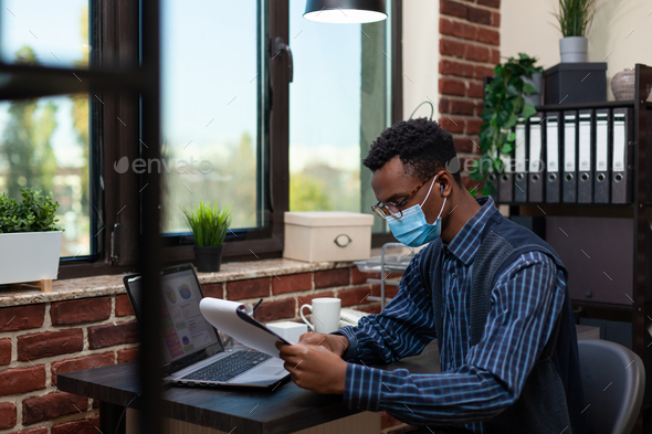Portrait of african american startup owner with glasses wearing face mask focused on clipboard