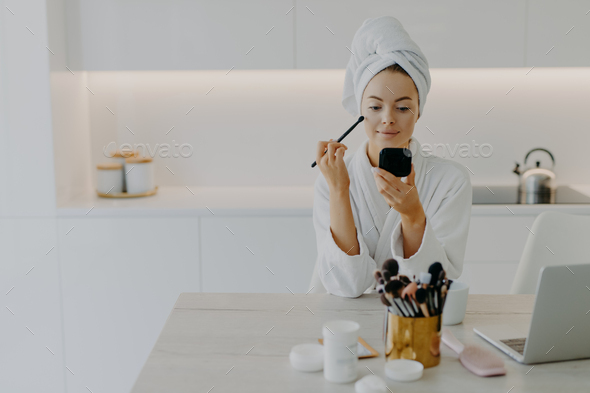 Refreshed lady applies face powder with cosmetic brush looks at herself in mirror poses at kitchen