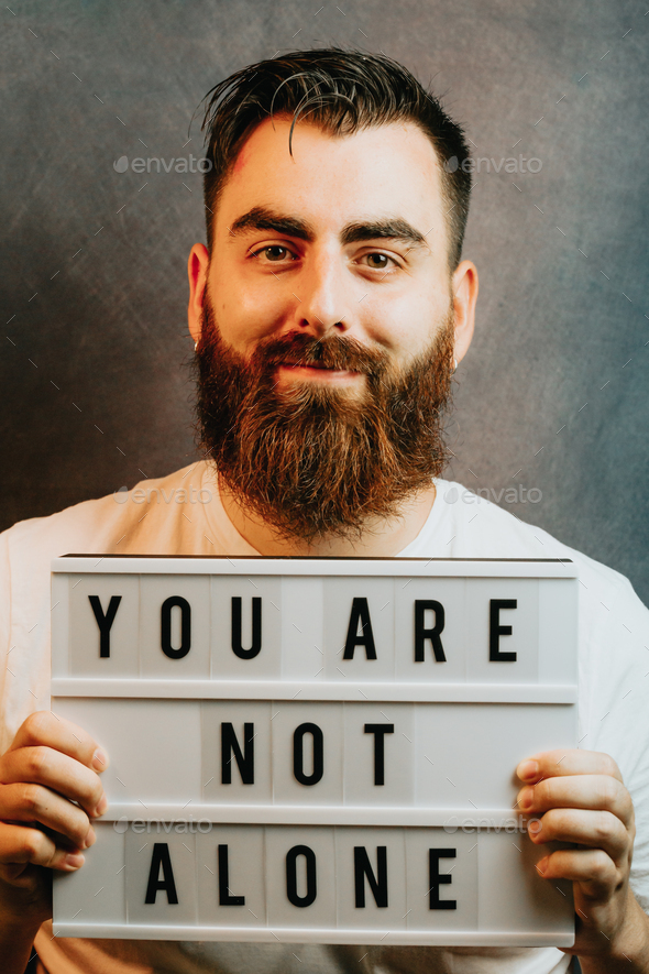 Young bearded hipster man holding a sign that says you are not alone, while smiling