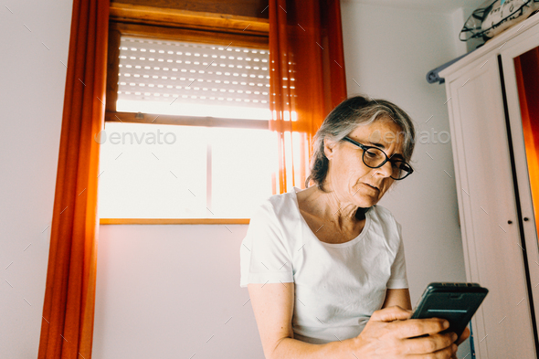 Old woman with glasses checking his phone and paying attention in his bedroom