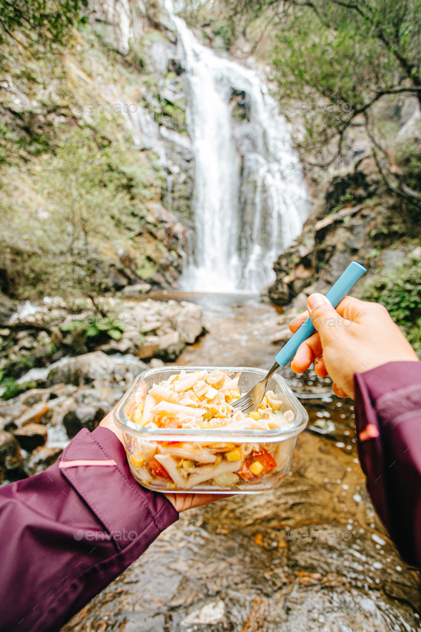 Point of view shot of a female hiker eating in front of a waterfall