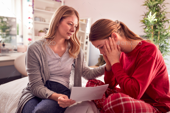 Disappointed Teenage Daughter Wearing Pyjamas Looking At School Exam Report With Mother In Bedroom