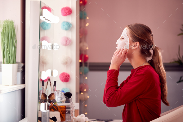 Teenage Girl Wearing Pyjamas Putting On Face Mask In Bedroom At Home