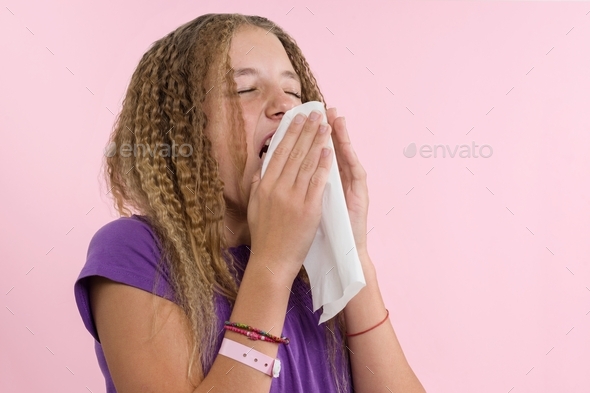 A girl in a T-shirt on a pink background sneezes into a napkin