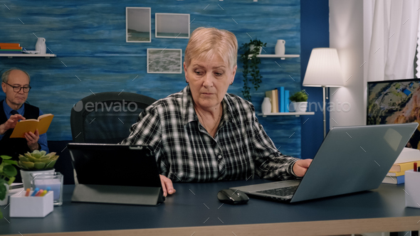 Old manager woman typing on laptop and reading on tablet in same time