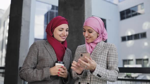 Two Young Muslim Women Wearing Hijab Headscarf Look at Phone, Walking Together Smile To Eash Other