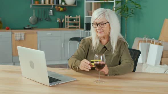 Happy Elderly Woman with a Glass of Wine Shopping Online From Home alt