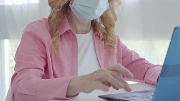 Unrecognizable Senior Woman in Face Mask Typing on Laptop Keyboard. Mid-adult Caucasian alt