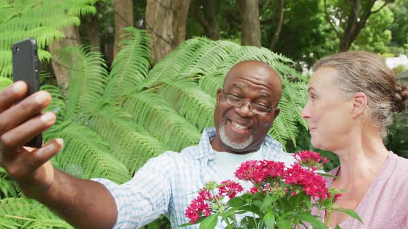 Happy senior diverse couple wearing shirts and taking selfie with smartphone in garden alt