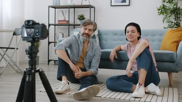 Mature Man and Teenage Boy Vloggers Recording Video with Camera at Home Showing Thumbsup and Waving alt