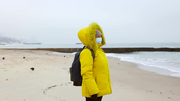 An Adult Woman in a Warm Yellow Jacket with a Backpack Uses a Protective Medical Mask Walking Walks