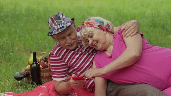 Family Weekend Picnic. Senior Old Grandparents Couple in Park Using Smartphone Online Browsing, Chat alt