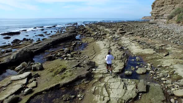 Tracking shot of a young man running on a rocky ocean beach shoreline alt
