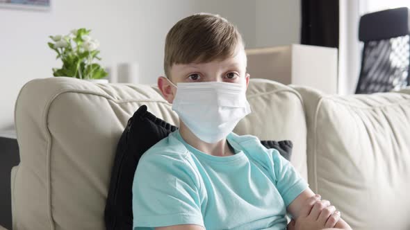 A Young Boy in a Face Mask Looks at the Camera As He Sits on a Couch at Home - Closeup alt