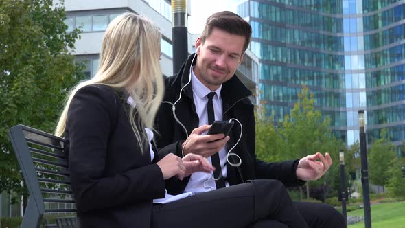 A Businessman and a Businesswoman Sit on a Bench and Listen To Music on a Smartphone in Urban Area alt