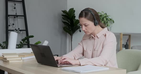 Smart Young Female Freelancer Wearing Headset Learning Distantly Sitting at Table with Books and alt