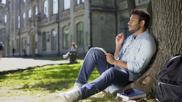 Multiracial Male Sitting on Grass Under Tree, Writing in Notebook, Creative Idea alt