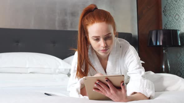 Young Redhead Woman Watching Movie on Tablet Computer Lying on Bed in Hotel Room