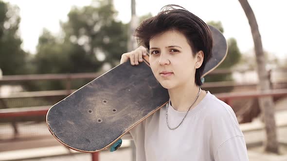 Teenage Girl Holding on the Shoulder Skateboard and Goes on Skatepark