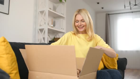 Stylish Middleaged Blonde Woman Sitting on the Couch with Carton Box on Laps alt