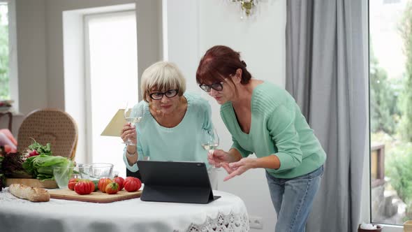 Two Confident Senior Female Friends Messaging Online on Tablet and Drinking White Wine Resting at alt