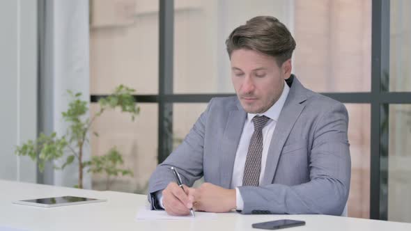 Middle Aged Man Writing on Paper in Office alt