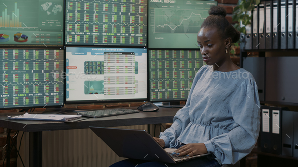 Female investor looking at real time stocks on laptop and multiple monitors - Stock Photo - Images
