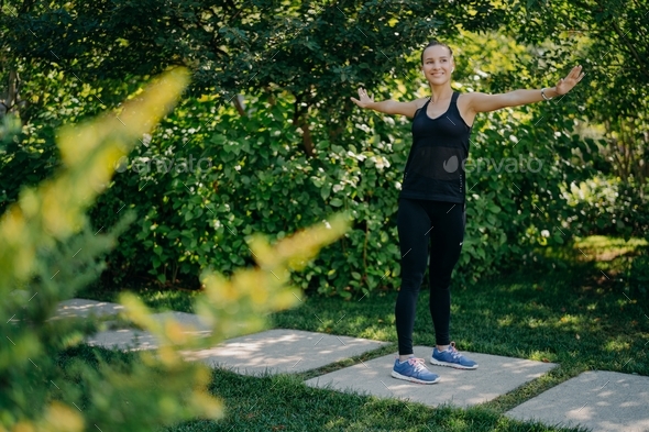 Athletic fit young woman keeps arms sideways being in good mood exercises outdoors
