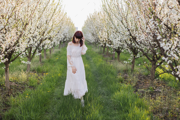 Gorgeous woman in white dress walking at blooming orchard