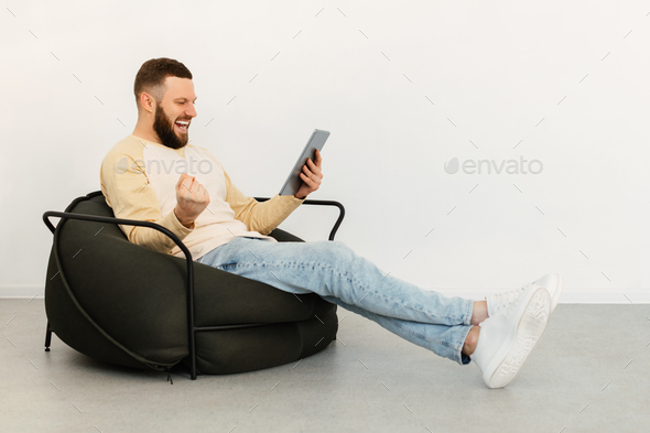 Man Using Digital Tablet Shaking Fists Sitting In Chair Indoors