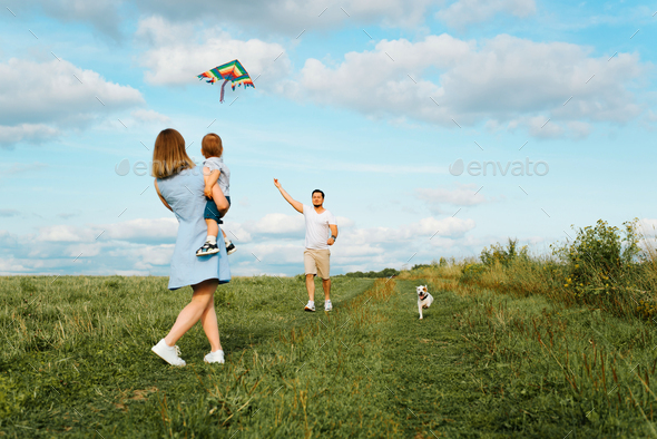 Happy family with child and dog playing in green field, smiling father runs with rainbow kite to