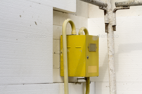 Close-up of yellow gas meter box with connecting pipes hanging on the corner of house wall insulated