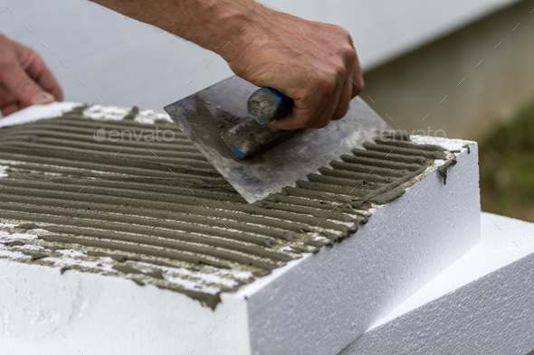 Close-up of worker hand with trowel applying glue on white rigid polyurethane foam sheet for house