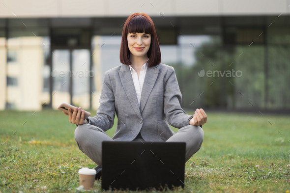 Calm young woman relaxing meditating, sitting outdoors on green grass with laptop