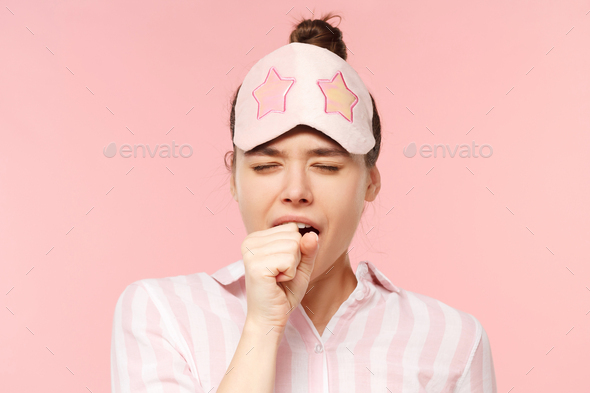 Girl in pyjama shirt and sleeping mask yawning and covering mouth, wanting to sleep