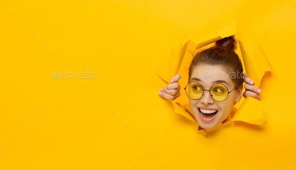 Young girl in glasses tearing paper and peeking out hole, curious about commercial offer