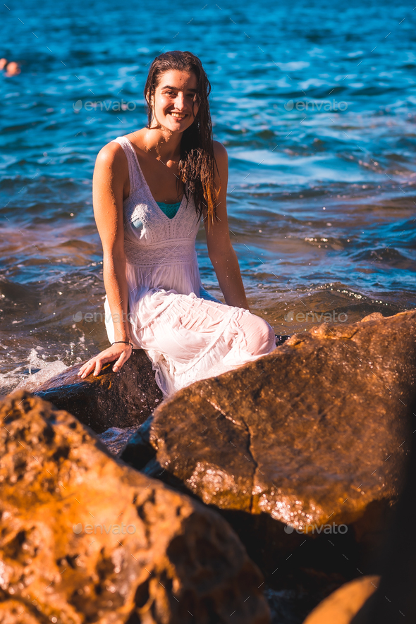 A young Caucasian woman in a wet white dress sitting on a rock Stock