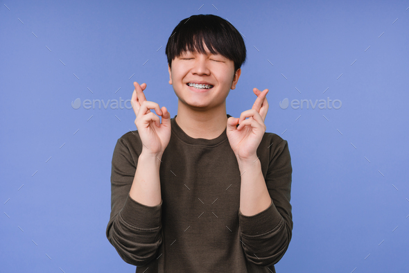Boy making a wish, crossing fingers for good luck before exams isolated in blue background