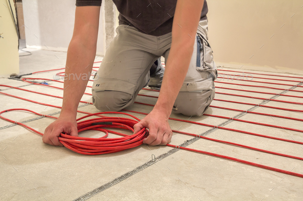 Electrician installing heating red electrical cable wire on cement floor in small new unfinished