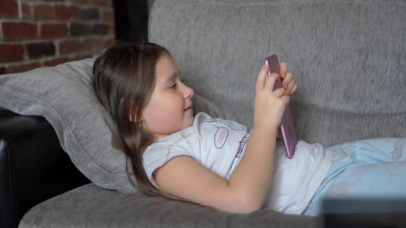 A young girl lying at home on the couch and looks into a pink tablet. She is funny and emotional