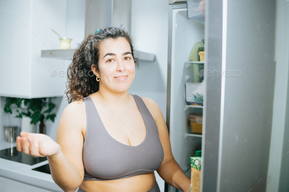 Curly woman doubting about what to eat from the fridge at the kitchen home to lose weight