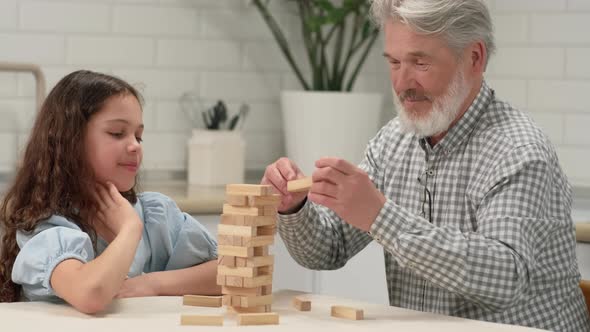 Grandfather and Granddaughter are Playing a Board Game at Home Removing Wooden Blocks From the Tower alt